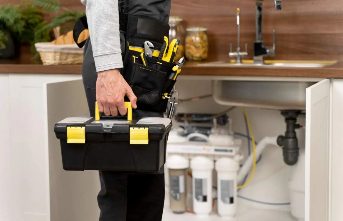 Person in work attire holding a toolbox, preparing to address plumbing or maintenance tasks under a sink.